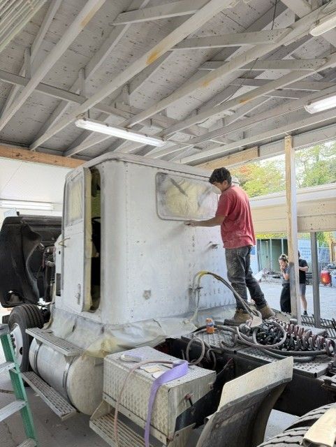 Worker spray-painting a white vehicle body inside a workshop, with tools and parts on the floor