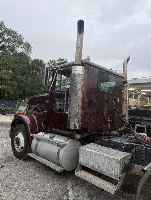 Old maroon semi truck with chrome exhaust and fuel tanks parked outdoors under overcast sky