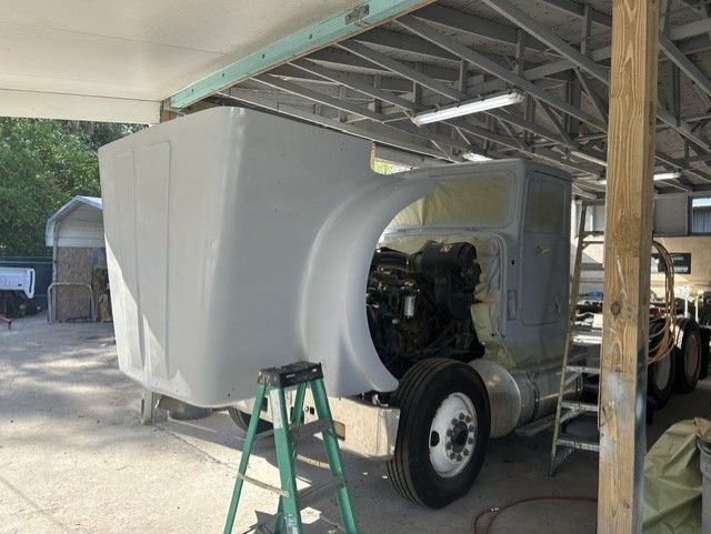 White truck cab shell under construction in a covered workshop, with exposed engine and tools nearby