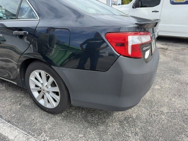 Rear side of a dark sedan with a gray rear bumper in a parking lot, showing minor damage near the wheel well