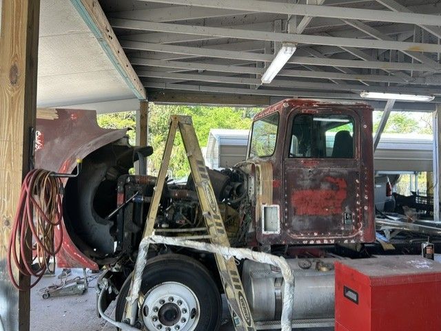 Rusty old truck under a shed with a red cabinet and large fan nearby, surrounded by tools and equipment