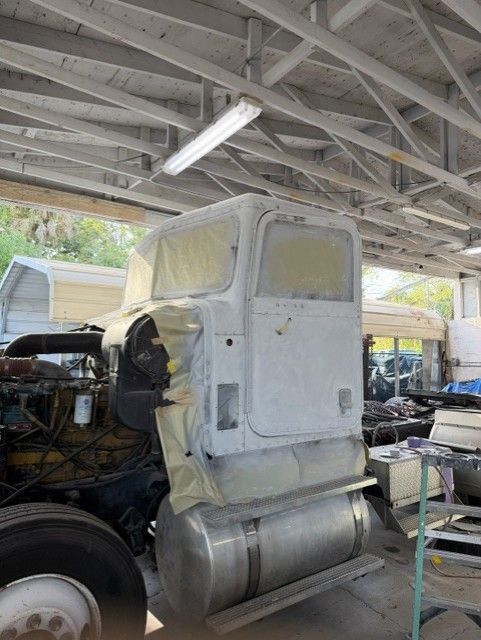 Old semi-truck cab in a workshop, partially painted white with exposed engine and silver fuel tank.