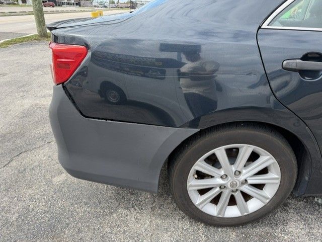 Rear side of a dark gray sedan with visible body damage near the left rear wheel in a parking lot
