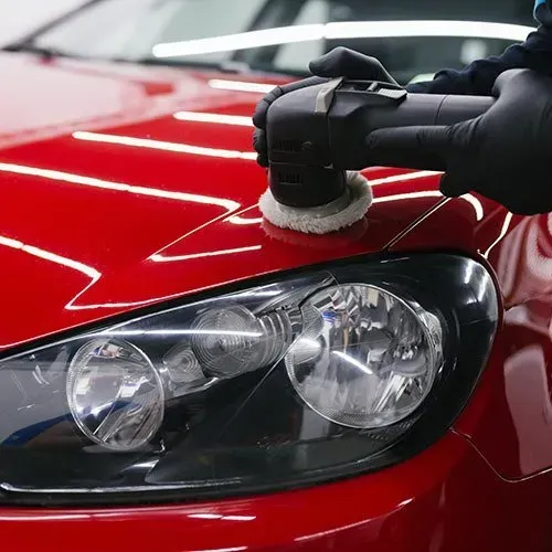 Person in black gloves polishing a red car with a buffer, close-up of headlight.