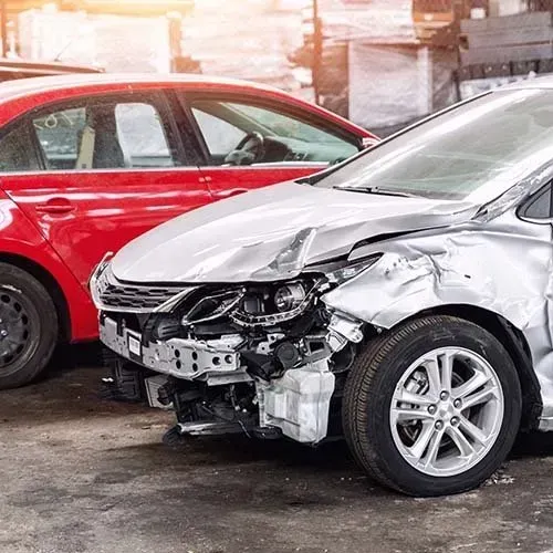 Damaged silver car with front-end damage, parked next to a red car. Cars appear to be in a repair shop.