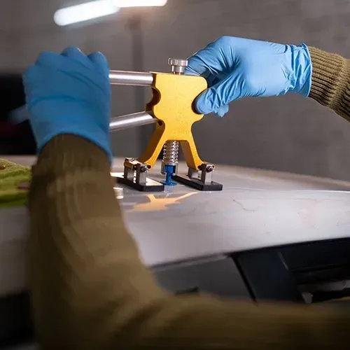 Person using a dent puller to repair a car. Hands wear blue gloves. Yellow tool on the car's white surface.