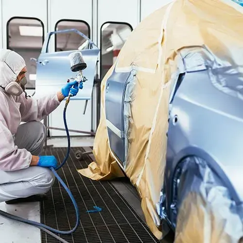 Person in protective suit painting a car with a spray gun in a body shop.