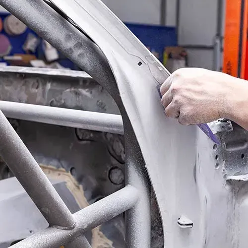 Person sanding a car door frame inside a workshop, covered in gray primer and a roll cage.
