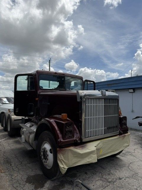 Vintage maroon semi truck parked outdoors with a chrome grille and covered bumper under a cloudy sky