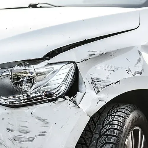 White car with front-end damage; crumpled hood, damaged headlight, and scratched fender.