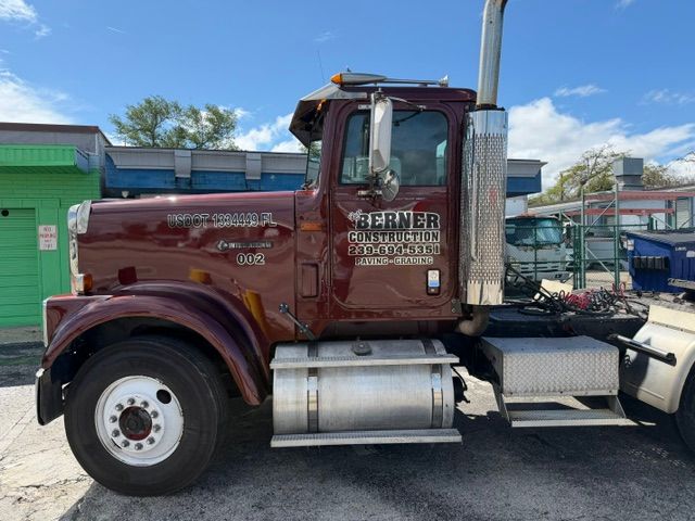 Maroon semi truck parked outdoors beside a green building under a blue sky