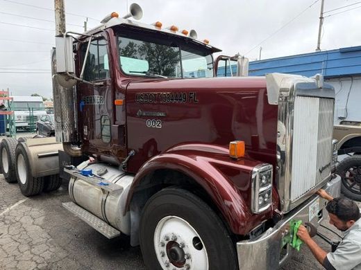 Maroon semi truck parked by a building while a worker kneels near the front bumper.