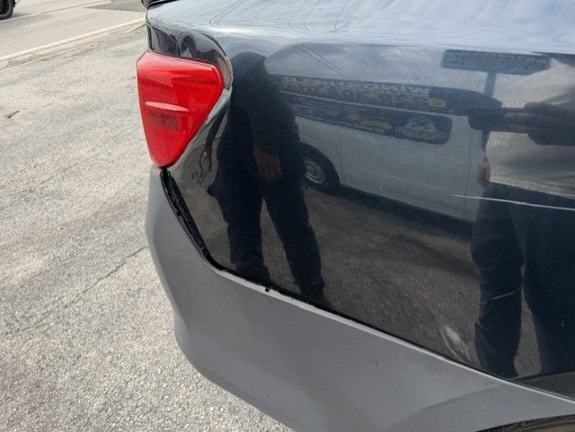 Close-up of a black car’s rear corner with a red taillight and gray bumper on a paved street
