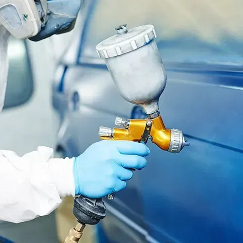 Person in protective gear sprays blue paint on a car with a spray gun.