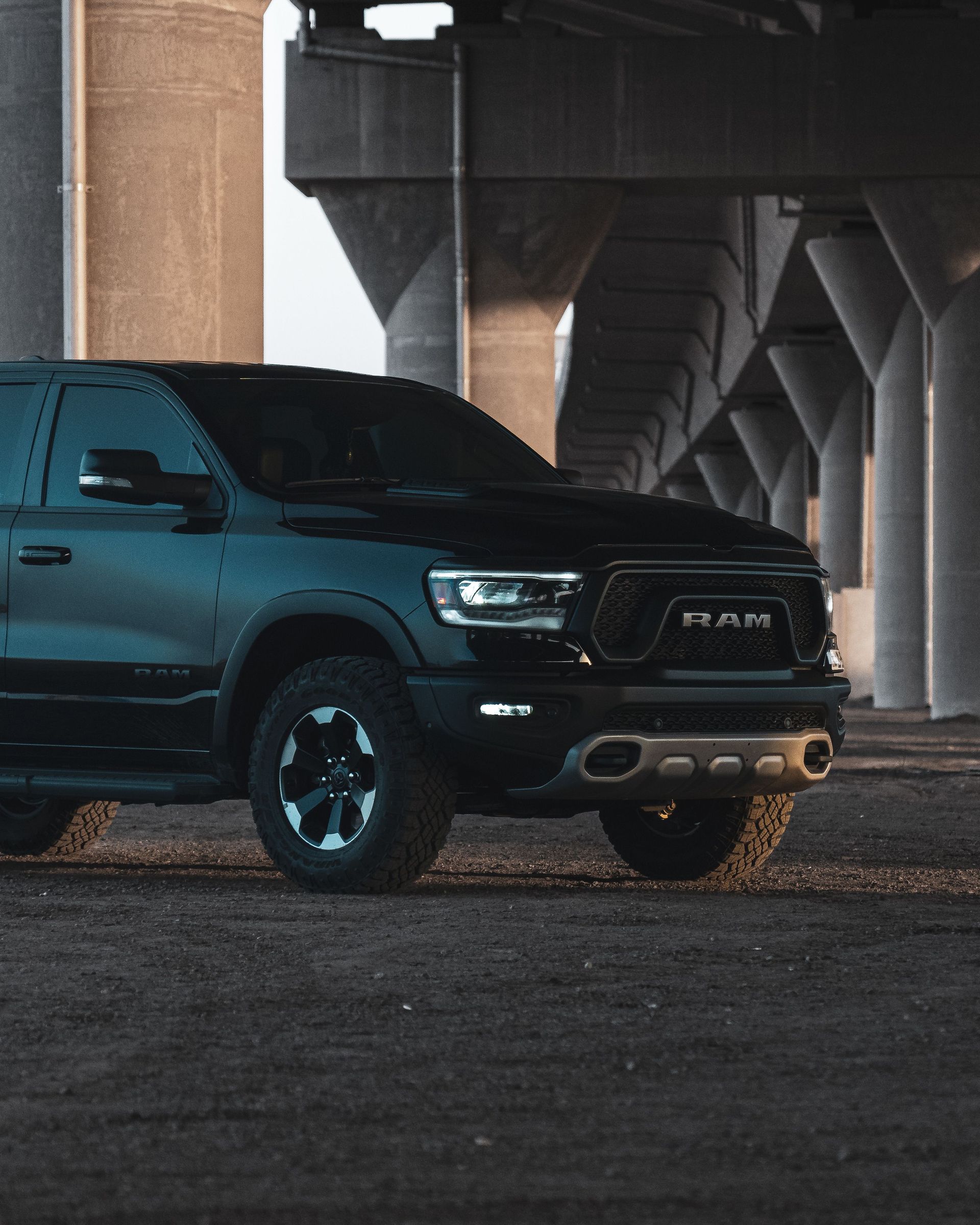 Black RAM pickup truck parked under a concrete overpass at dusk.
