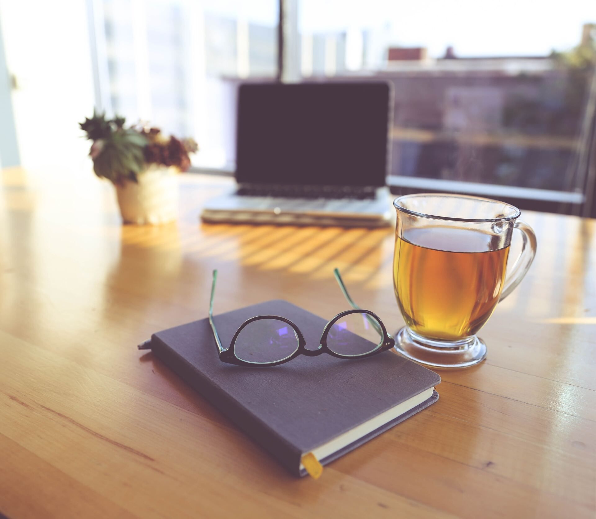 Notebook, eyeglasses, and tea on a wooden desk beside a laptop in soft sunlight.
