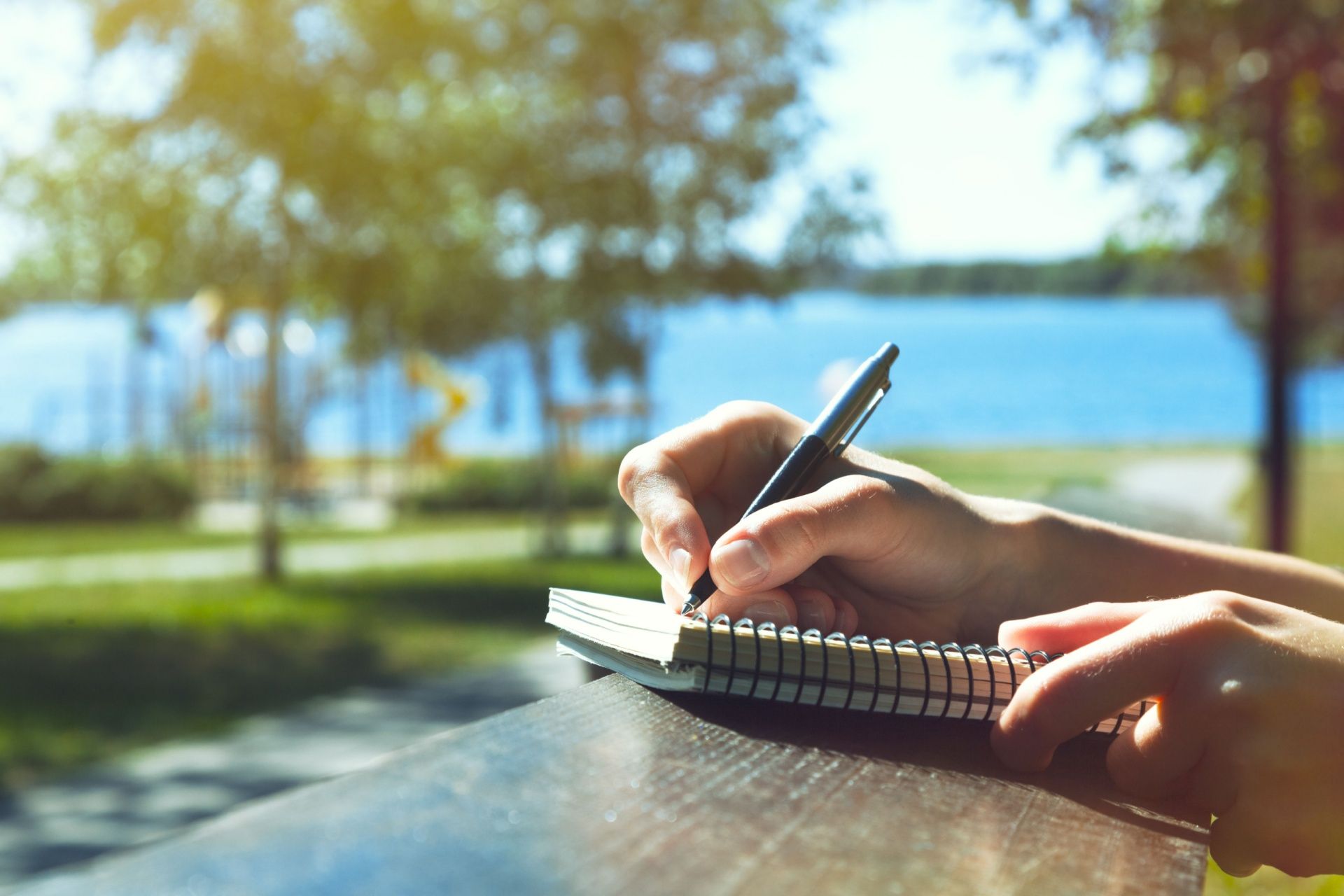 Hand writing in a spiral notebook by a sunny lakeside park path