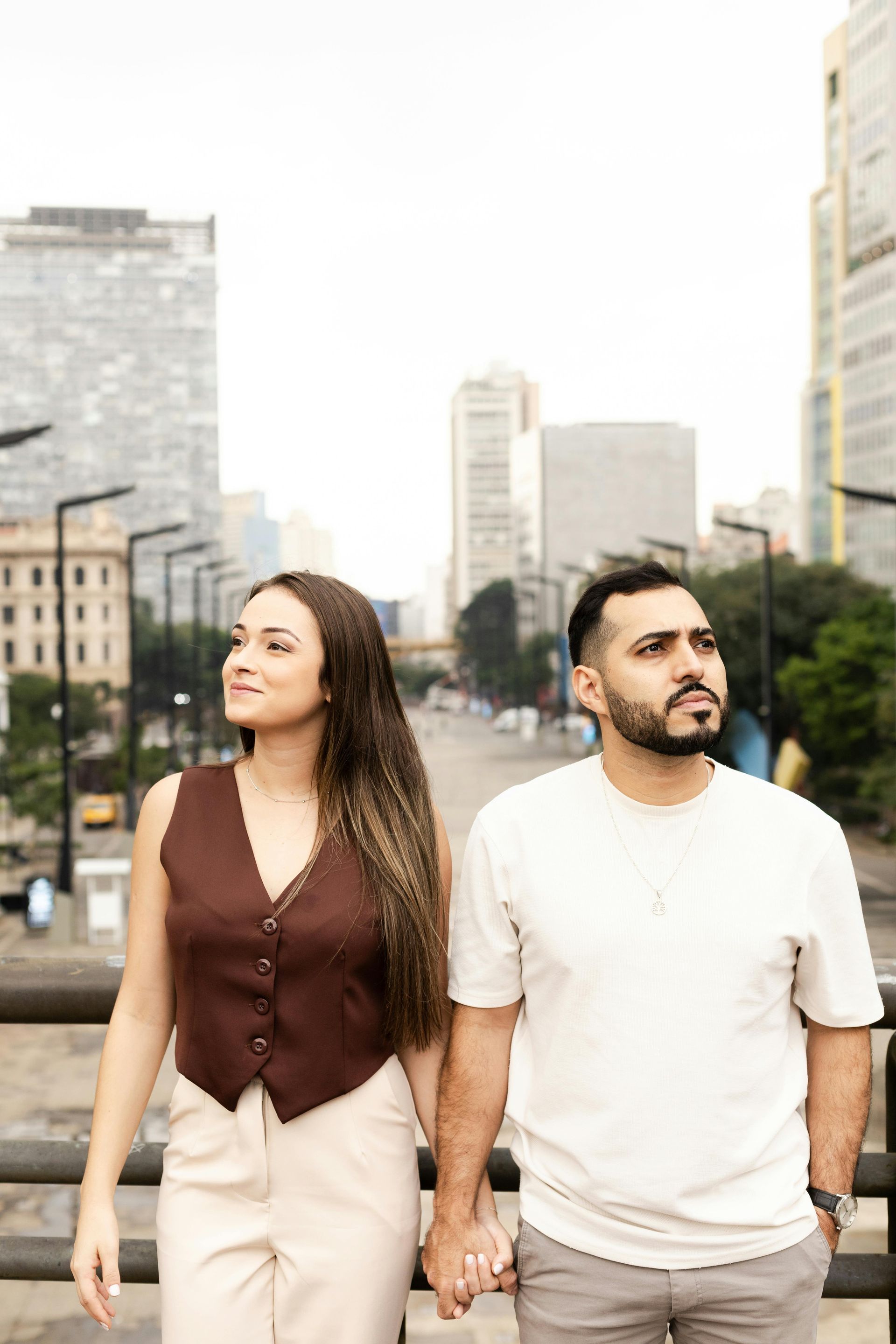 Couple holding hands and walking in a city street, looking off to the side, with tall buildings behind them.