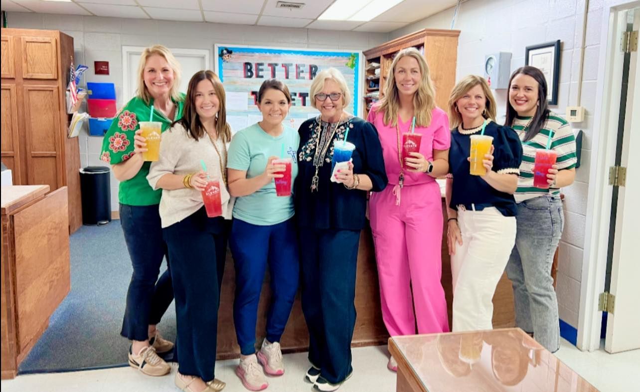 Seven people stand in a line indoors, holding colorful drinks and smiling in front of a sign that says 