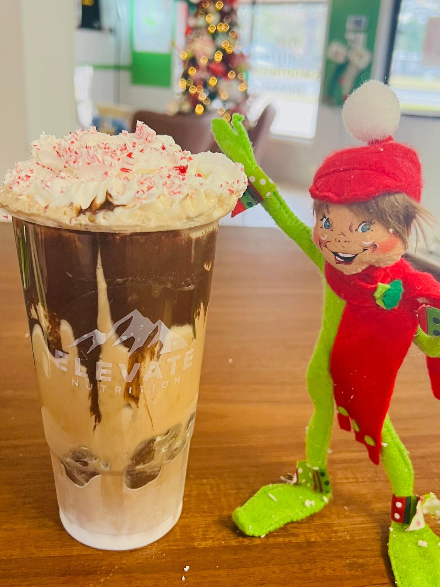 A festive holiday-themed drink in a clear plastic cup topped with whipped cream next to a small, cheerful elf doll.