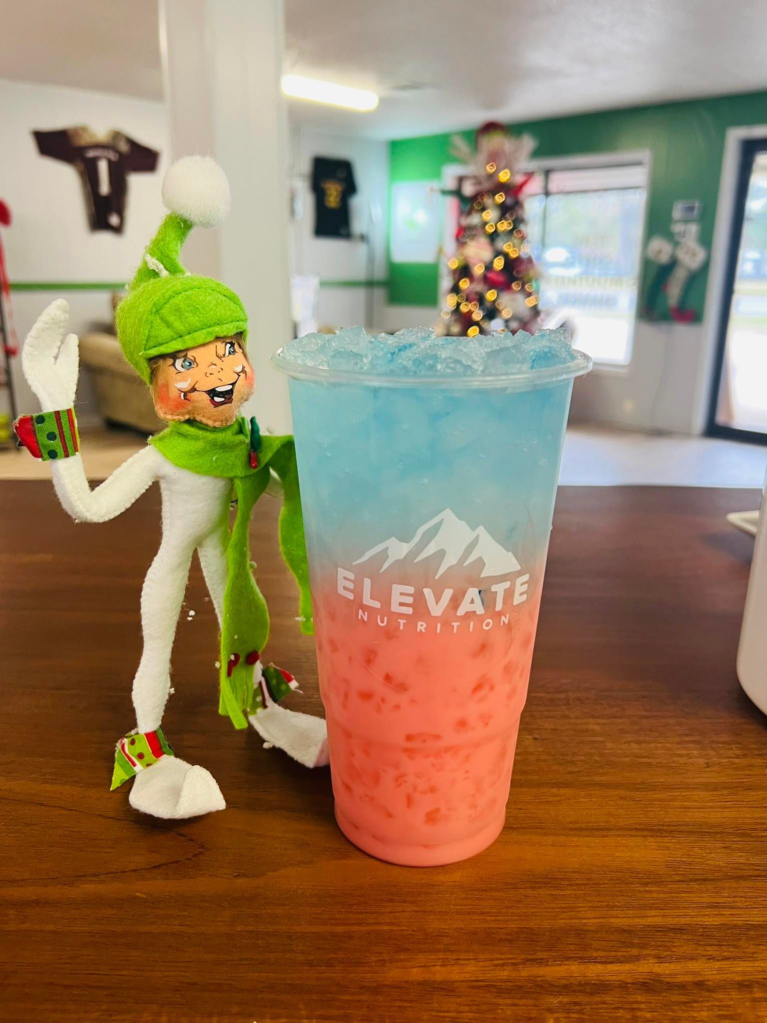 A green-clad elf figurine stands next to a blue and pink gradient drink in a cup labeled 