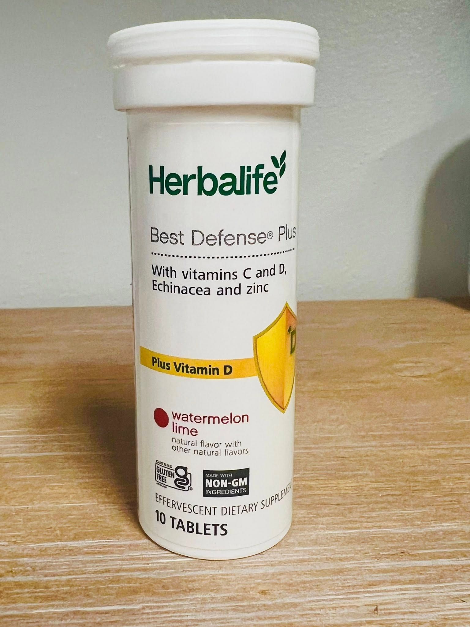 A white cylindrical bottle of Herbalife Best Defense Plus tablets with a yellow shield icon on a wooden surface.