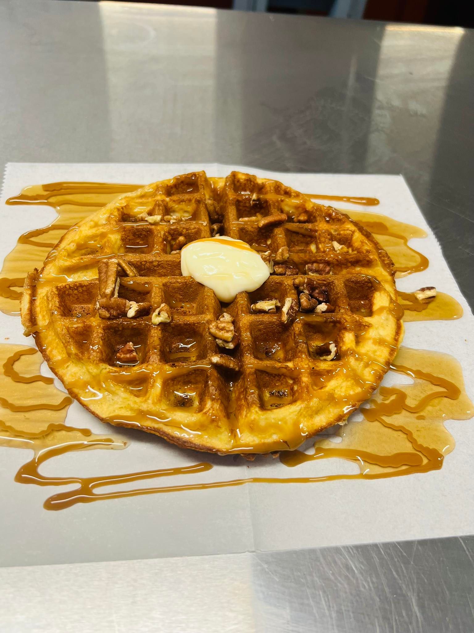 A golden-brown Belgian waffle topped with pecans and a pat of butter, drizzled with syrup on white parchment paper.