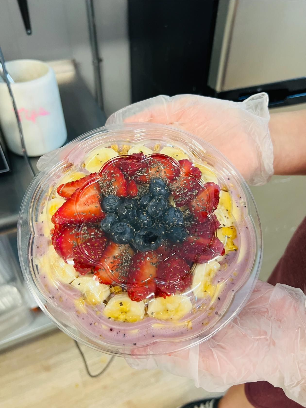A gloved hand holds a clear plastic bowl containing a smoothie topped with sliced bananas, strawberries, and blueberries.