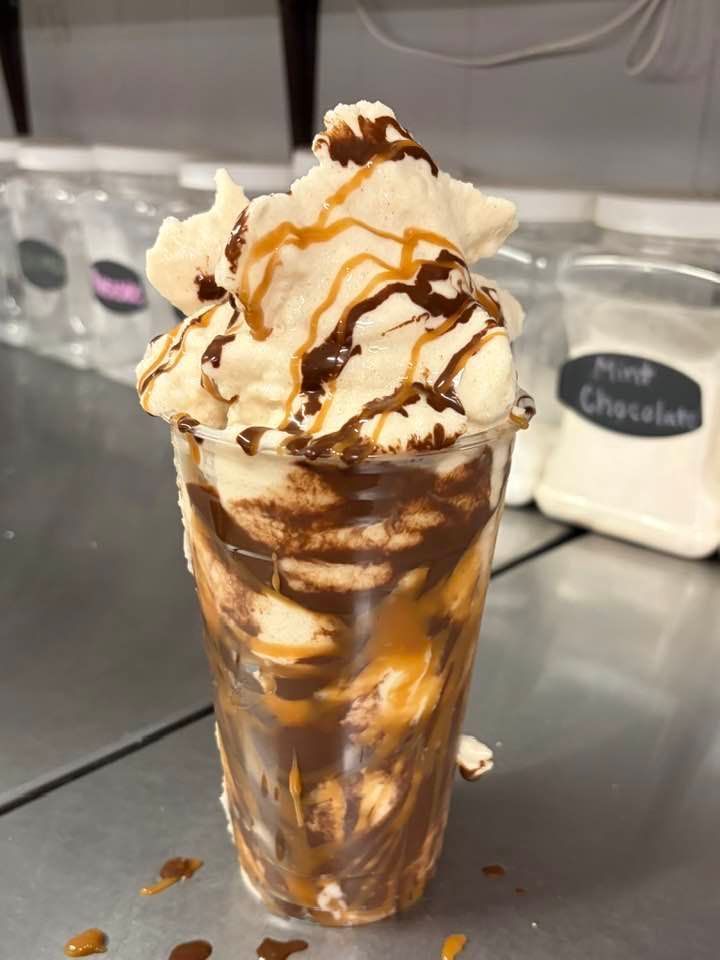 A clear plastic cup filled with a chocolate and caramel milkshake, topped with whipped cream, chocolate, and caramel sauce.