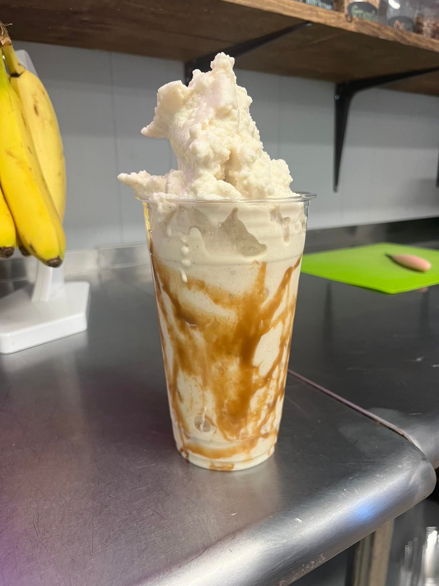 A smoothie topped with a scoop of frozen yogurt, drizzled with caramel sauce in a clear cup, with bananas nearby.