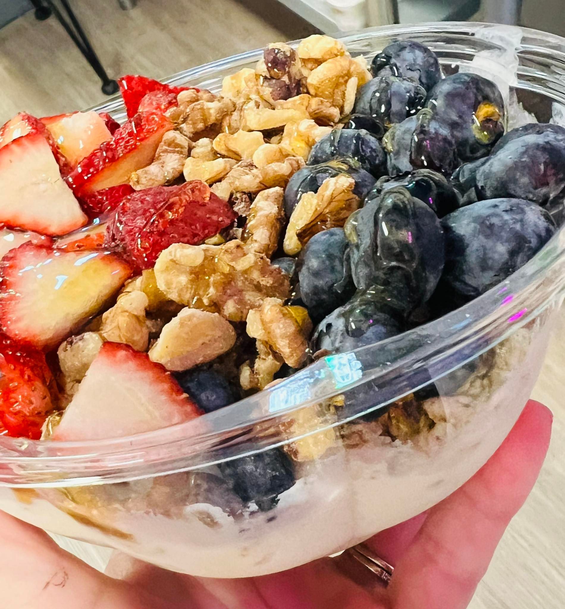 A hand holds a plastic bowl filled with yogurt, topped with sliced strawberries, blueberries, and crunchy granola.
