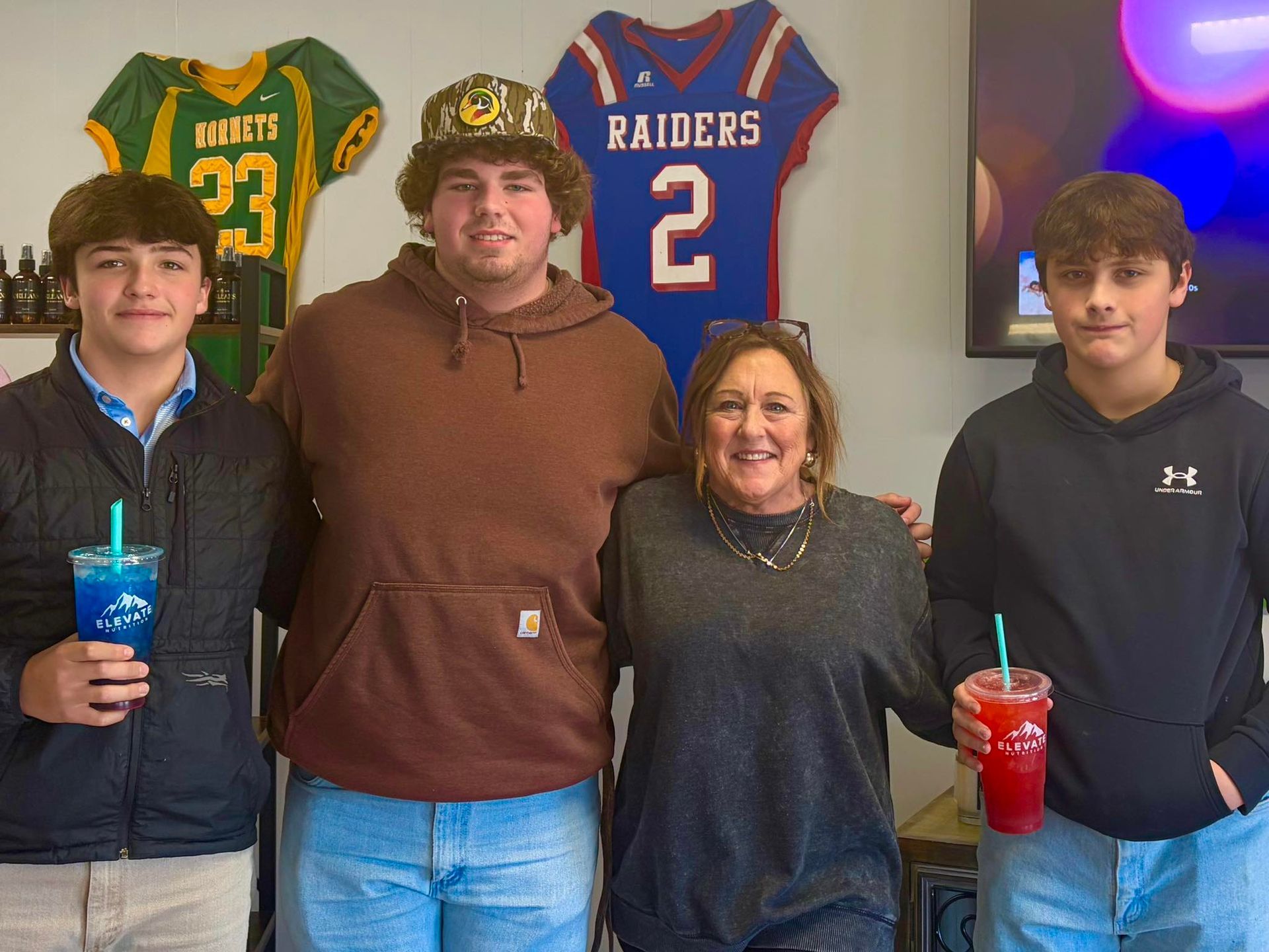 Four people posing for a photo indoors in front of sports jerseys; two individuals are holding colorful drinks.