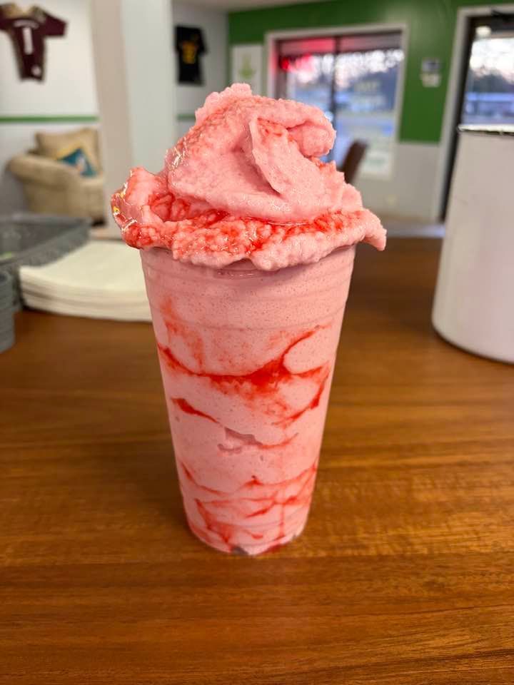 A tall, pink strawberry smoothie with whipped topping, sitting on a wooden counter in a shop.