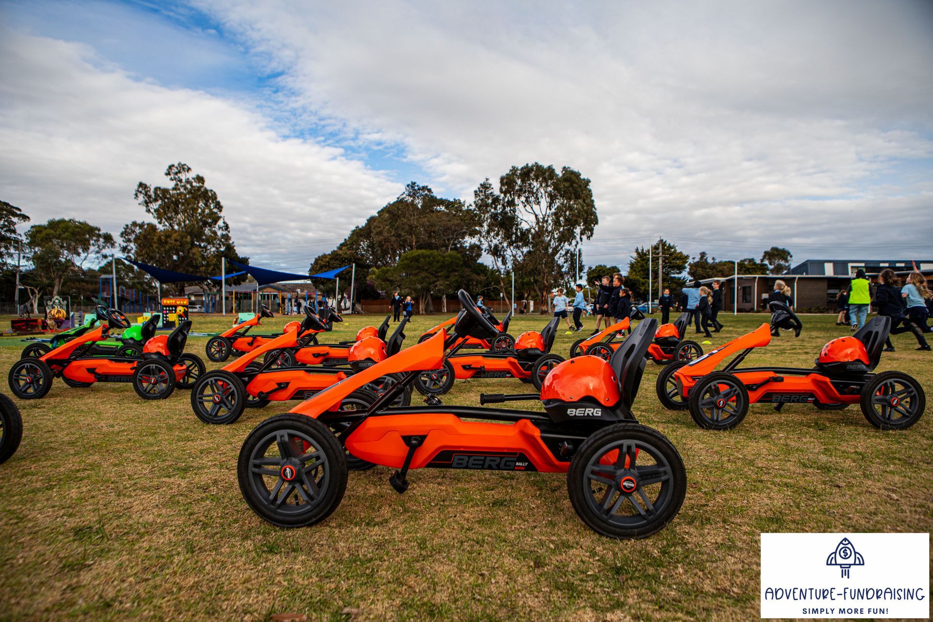 Orange pedal karts on grass field, ready for an event. People in the background.