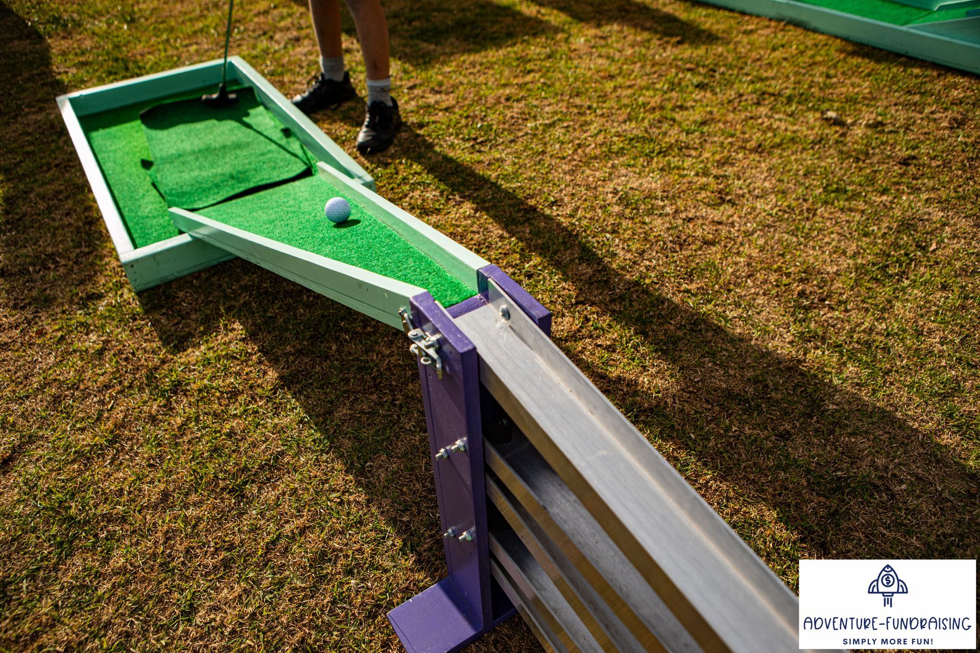 Miniature golf setup with green turf, ball, and ramp leading to a silver and purple track on grass.