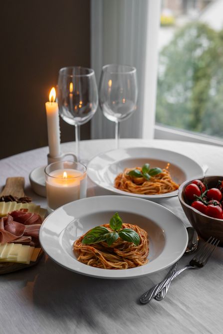 Romantic dinner table with pasta, wine glasses, candles, and berries by a window