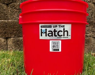 Red plastic bucket with “Up the Hatch” label sitting on grass in front of a stone wall