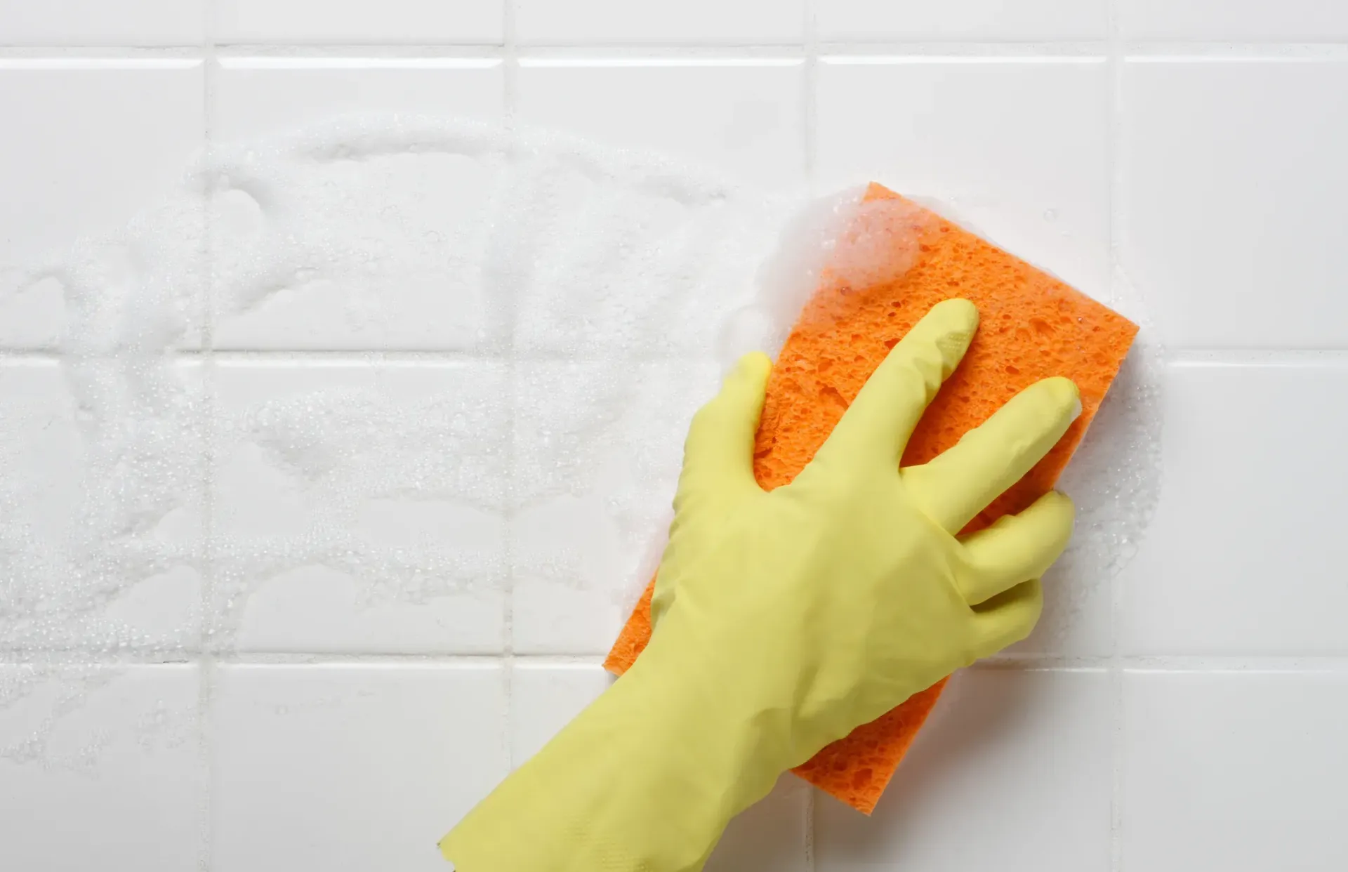 A person wearing yellow gloves is cleaning a tile wall with a sponge.