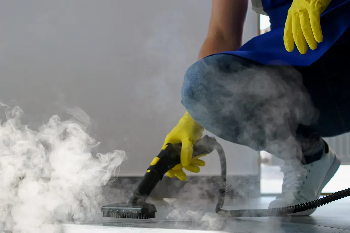A person wearing yellow gloves is using a steam cleaner to clean a floor.