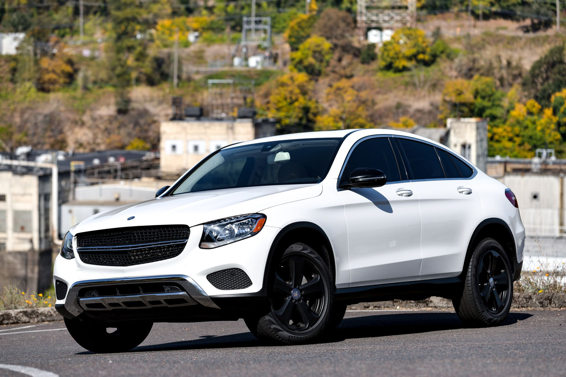 White Mercedes-Benz SUV on a road, black wheels, against a backdrop of buildings and trees.