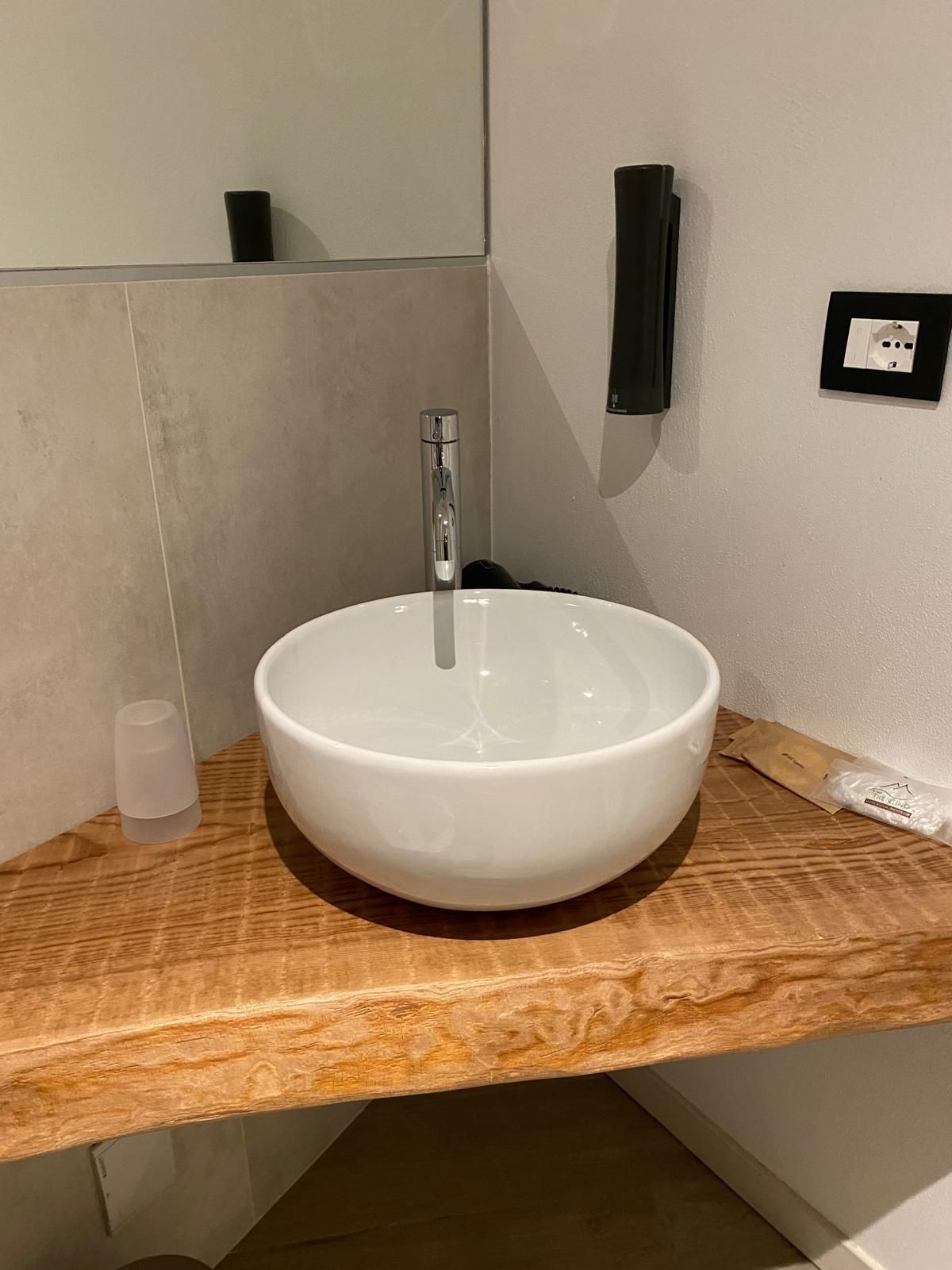 Lavabo di piccole dimensioni con rubinetto cromato su un piano in vimini, accanto a uno specchio e a un dispenser a parete.