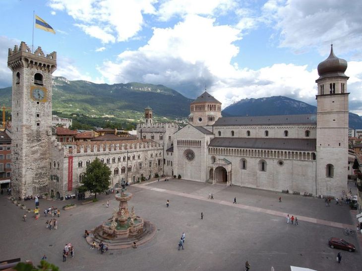 Piazza storica in pietra con cattedrale e torre dell'orologio, montagne sullo sfondo e una fontana nella piazza