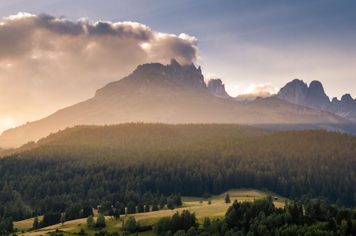 Cresta montuosa illuminata dal sole sopra una valle boscosa all'alba, con le nuvole che fluttuano nel cielo.
