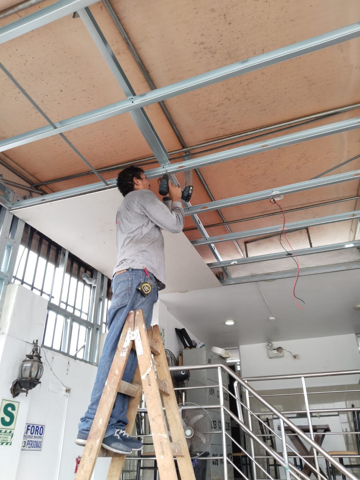 Trabajador usando un taladro mientras está subido a una escalera, instalando la estructura del techo en un espacio interior.