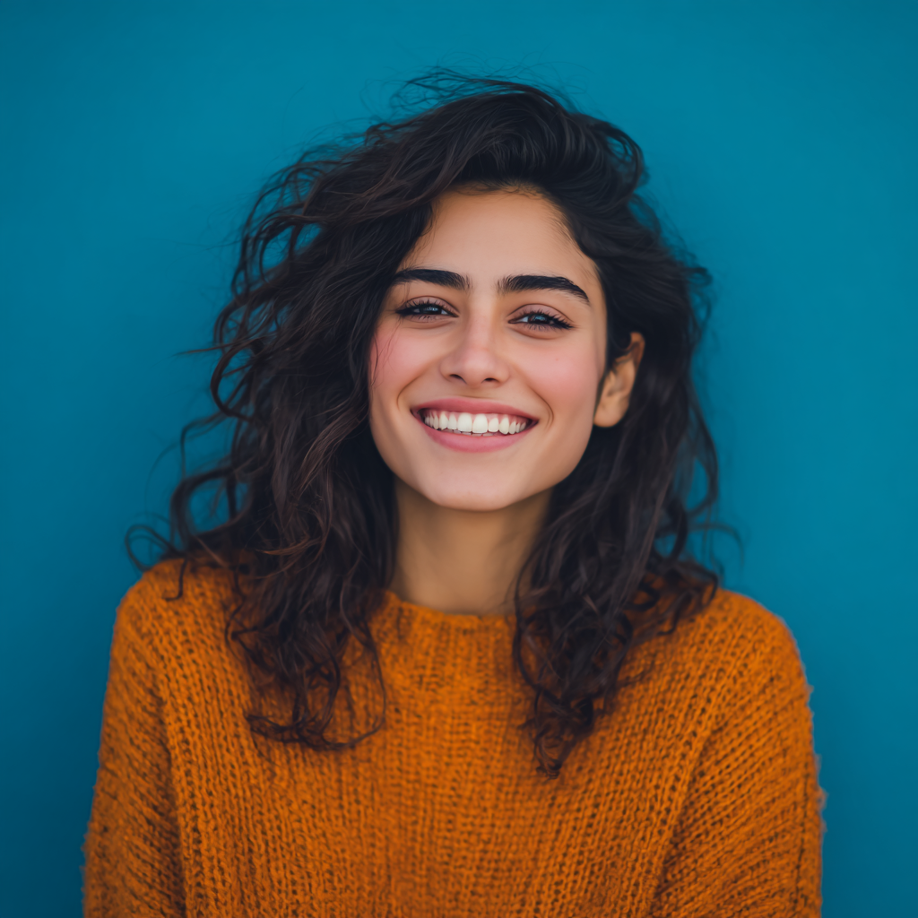 Woman with dark curly hair and a wide smile, wearing an orange sweater against a blue background.