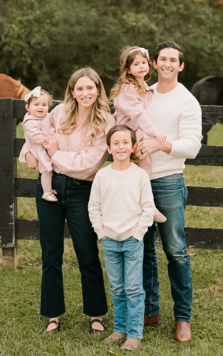 Family of five posing outdoors in a field, smiling and dressed in light sweaters and jeans.