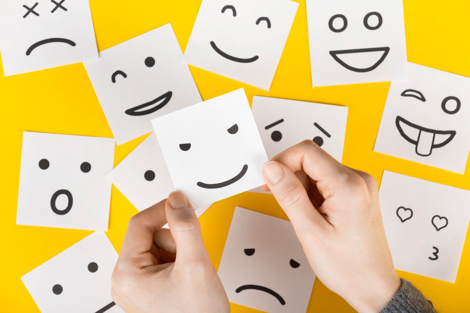 Hands holding a neutral smiley face card among other illustrated emotions on yellow background.
