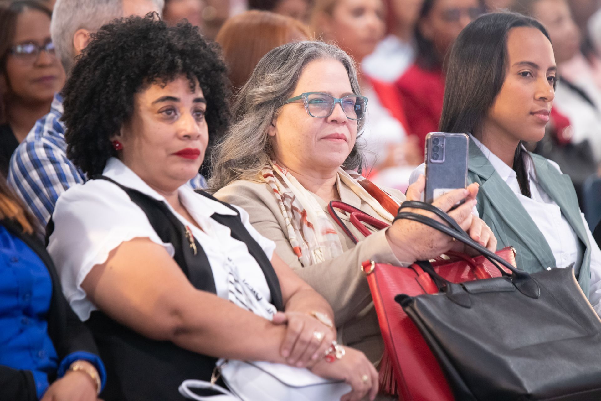 Three women seated, one holding a phone. Middle woman wearing glasses and a scarf, other two look on.