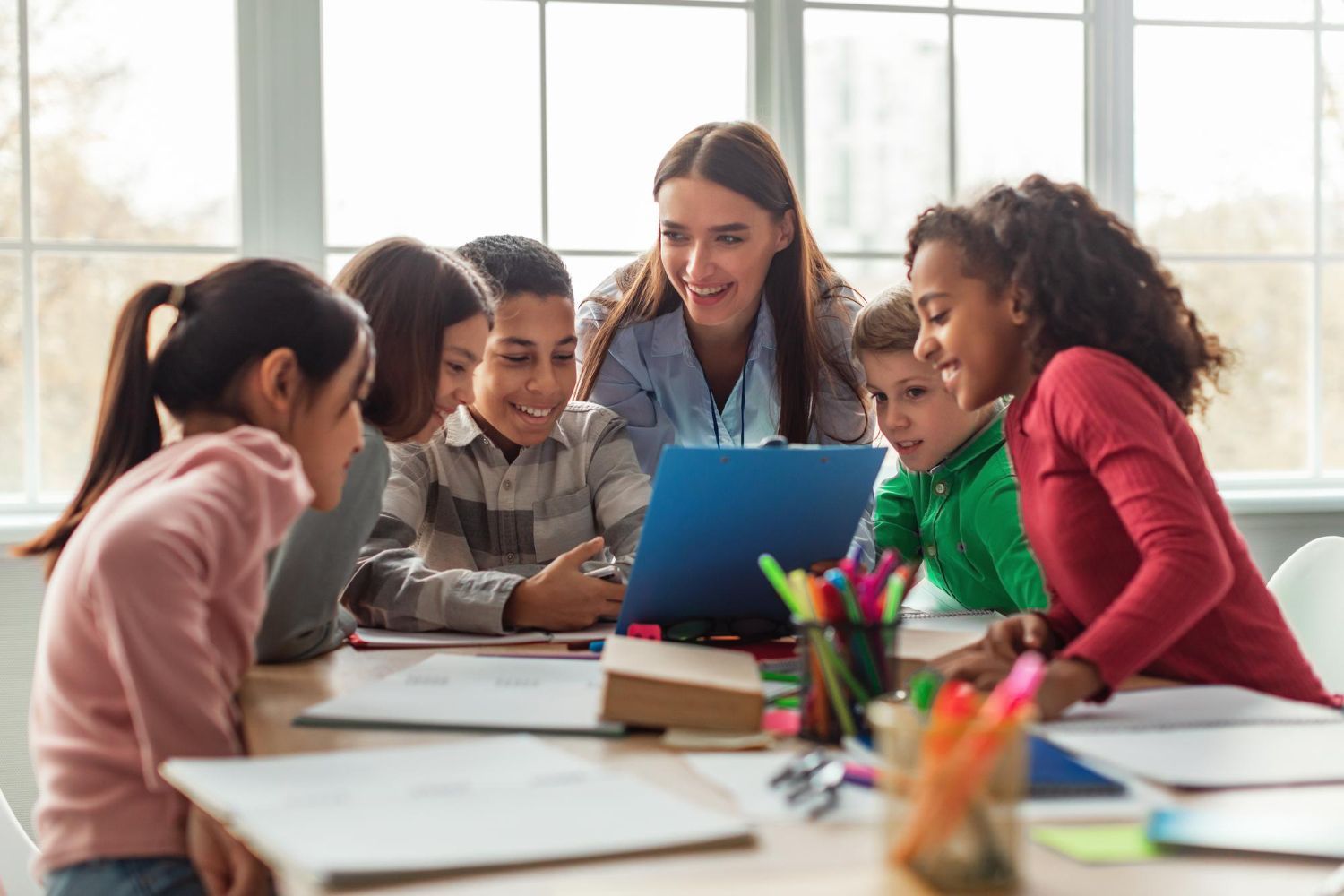 Teacher and students looking at a laptop together at a desk. Smiling. Classroom setting.