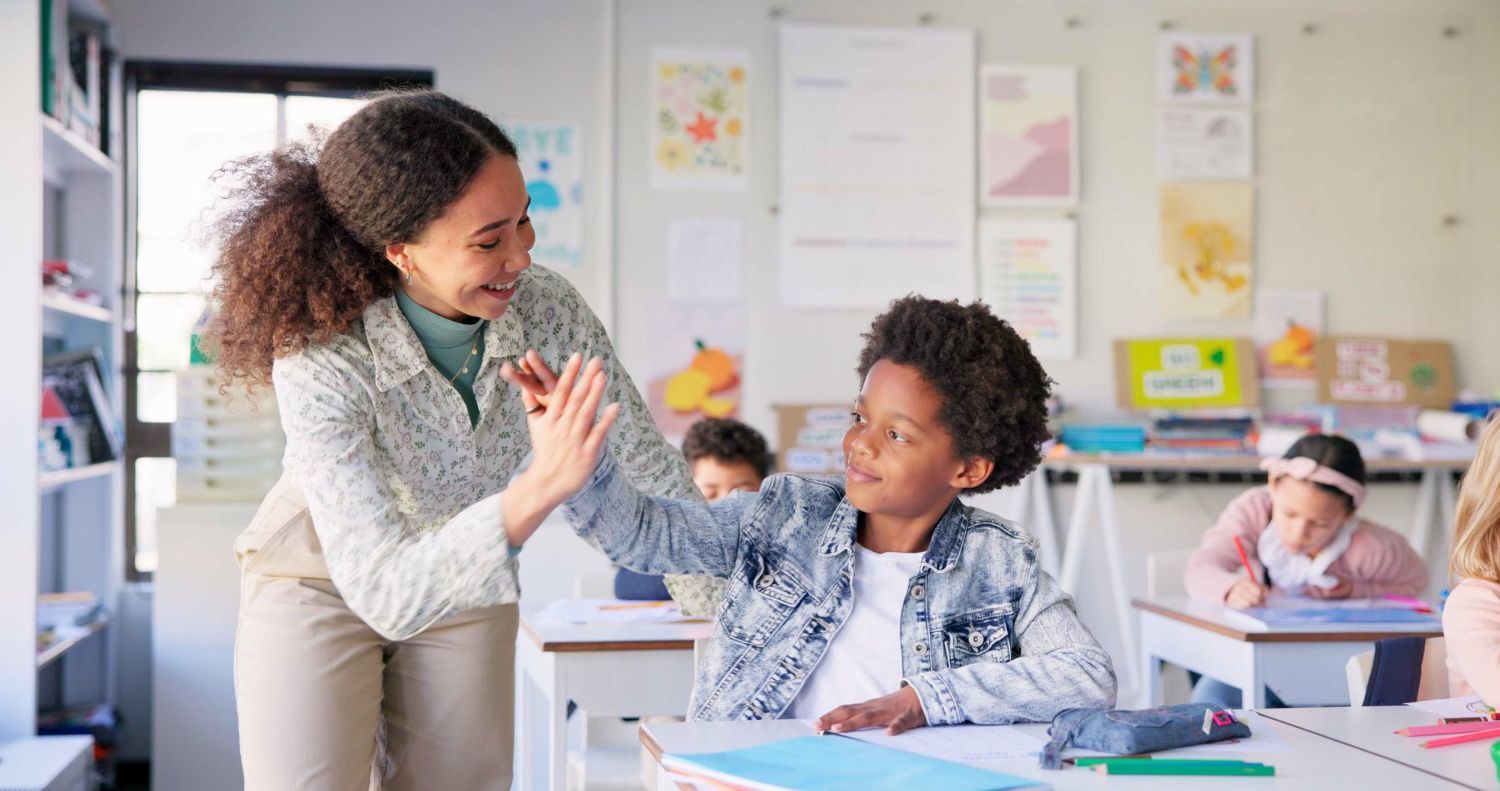 Teacher high-fives a student at their desk in a classroom. Other students are seated at desks.